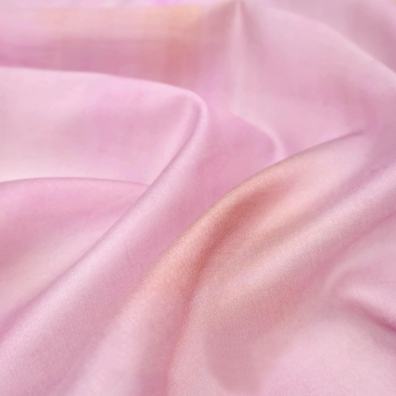 Op de afbeelding: Close-up van een vloeiende, lichtroze stof met subtiele kleurvariaties. De stof heeft zachte plooien en een gladde textuur, waardoor een zachte, etherische uitstraling ontstaat. De kleur gaat over van een zachtroze naar een warmere, perzikkleurige tint.