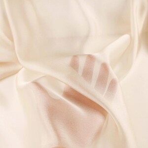 May include: Close-up of a flowing, cream-coloured fabric. The fabric is translucent, with a hand visible beneath the material. The light catches the folds, creating highlights and shadows. The fabric appears soft and delicate, possibly silk or a similar material.