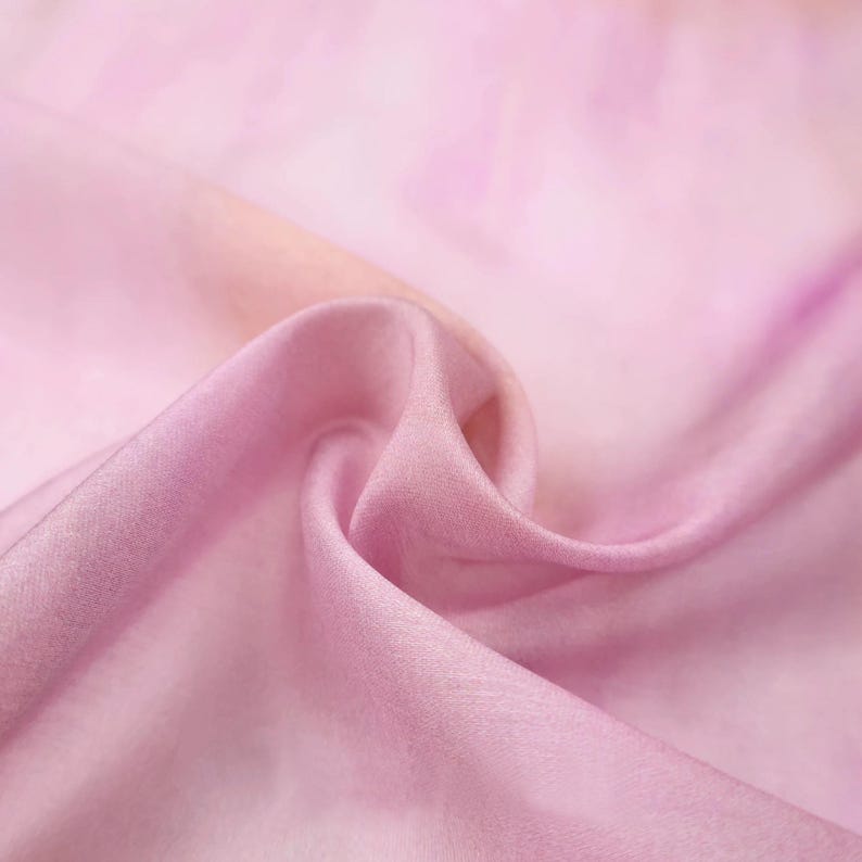 Op de afbeelding: Close-up van een zachte, vloeiende stof in roze en perziktinten. Het materiaal lijkt zijde of een vergelijkbaar delicaat textiel te zijn, met subtiele variaties in kleur en textuur. De stof is gedrapeerd, waardoor zachte plooien en schaduwen ontstaan.