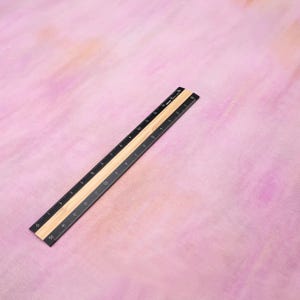 Op de afbeelding: Een zwart en bruin liniaal met inch- en centimeter markeringen, schuin op een roze en perzik aquarel achtergrond. De liniaal is gemaakt van hout en metaal, met cijfers en lijnen voor precieze metingen.