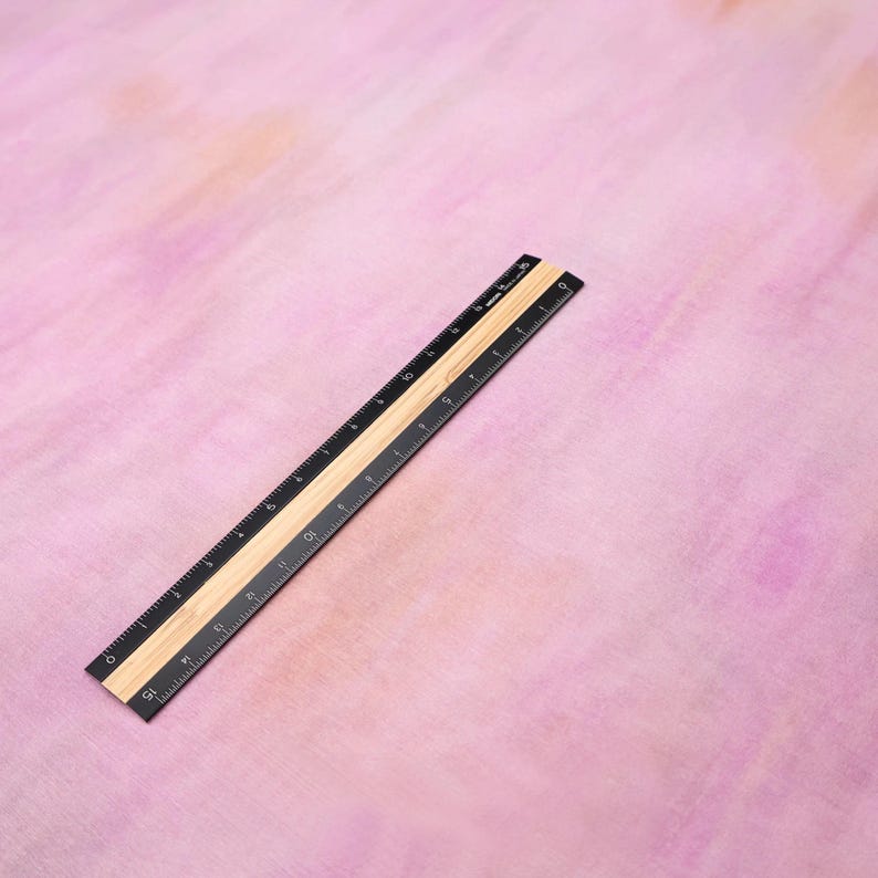 Op de afbeelding: Een zwart en bruin liniaal met inch- en centimeter markeringen, schuin op een roze en perzik aquarel achtergrond. De liniaal is gemaakt van hout en metaal, met cijfers en lijnen voor precieze metingen.