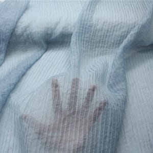 May include: Light blue sheer fabric with a textured, pleated design. The fabric is draped, with a hand visible through the material, creating a sense of depth and transparency. The fabric has a soft, flowing appearance.
