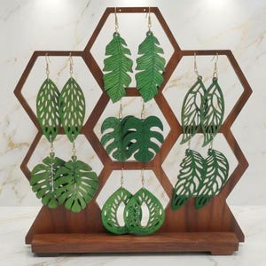 May include: A wooden honeycomb display stand holding five pairs of green leaf-shaped earrings. The earrings feature various botanical cut-out designs, including long slender leaves, monstera leaves, and teardrop shapes with intricate patterns.