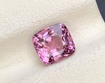 Op de afbeelding: Een gefacetteerde, kussenvormige edelsteen in een zachte roze tint. De edelsteen ligt op een wit oppervlak, met een donkere achtergrond. De facetten van de steen reflecteren licht, waardoor een sprankelend effect ontstaat. Deze edelsteen is waarschijnlijk voor sieraden.