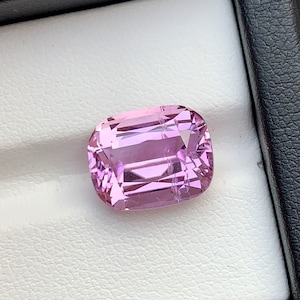 May include: A cushion-cut, light pink gemstone rests in a black jewelry box. The gemstone has multiple facets that reflect light, creating a sparkling effect. The gem is a soft, pastel pink color.