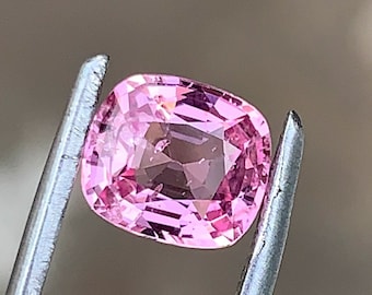 Op de afbeelding: Een roze, kussenvormige edelsteen wordt vastgehouden door een zilveren pincet. De edelsteen heeft een vierkante vorm met afgeronde hoeken en een gefacetteerd oppervlak. De edelsteen heeft een lichte, levendige roze kleur.