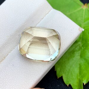 以下が含まれることがあります： 正方形のクッションカットが施された、薄い黄色のファセット宝石。 宝石は白い表面に置かれ、背景には緑の葉があります。 宝石のファセットは光を反射し、きらめく効果を生み出しています。