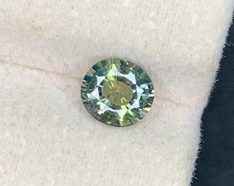 May include: A round, faceted gemstone with a mix of green and yellow hues. The gem is set against a white background. The facets create a starburst pattern in the center.