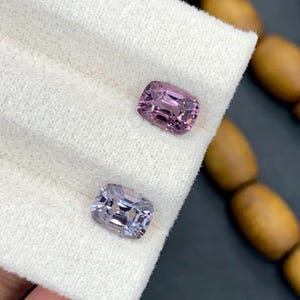 Può includere: Due gemme taglio cuscino, nelle tonalità del viola e lavanda, poggiano su un panno bianco. Le gemme hanno molteplici sfaccettature, che riflettono la luce. Lo sfondo include perline di legno e una superficie scura.