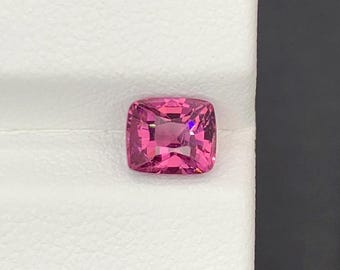 May include: A cushion-cut, vibrant pink gemstone. The gem is set against a white background, with a dark gray edge. The gemstone's facets reflect light, creating a sparkling effect. This gem could be used in jewelry.