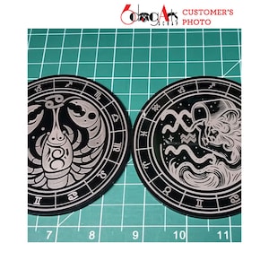 May include: Two black coasters with white zodiac signs. The left coaster features a crab with the sign Cancer. The right coaster features a person pouring water with the sign Aquarius.
