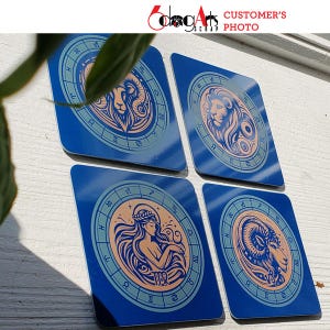May include: Four blue square coasters with a zodiac sign design. Each coaster features a different zodiac sign with a gold and peach colored illustration. The signs are Aries, Leo, Virgo, and Capricorn.