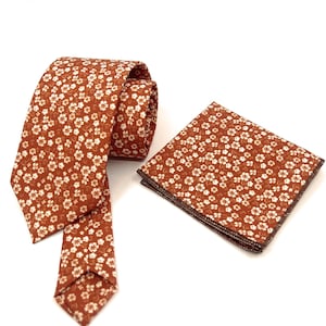 May include: A necktie and a matching square pocket handkerchief, both featuring a burnt orange background with a repeating pattern of small white five-petaled flowers. The necktie is partially rolled, and the pocket square is folded flat.