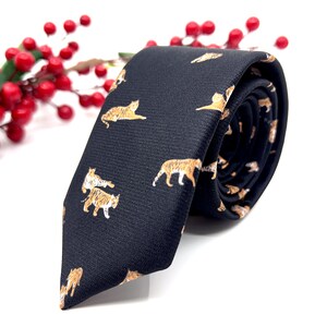 May include: A black necktie featuring a repeating pattern of small orange and white tigers in various poses. The fabric has a subtle textured weave, and the tie is rolled at the end, displaying the printed tiger motifs across the dark surface.