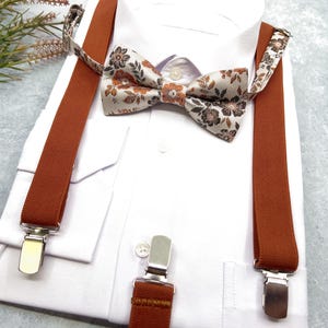 May include: A white dress shirt paired with a set of solid burnt orange suspenders and a matching bow tie. The bow tie features a detailed floral pattern in shades of brown, orange, and cream. The suspenders include silver metal clips.