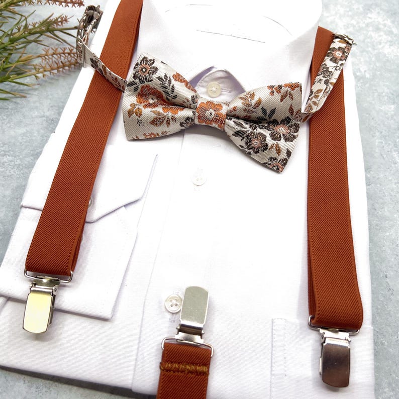 May include: A white dress shirt paired with a set of solid burnt orange suspenders and a matching bow tie. The bow tie features a detailed floral pattern in shades of brown, orange, and cream. The suspenders include silver metal clips.