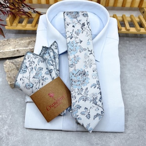 May include: A light blue dress shirt paired with a matching necktie and pocket square featuring a floral pattern in shades of blue and grey. A brown paper tag with the text Cranebird Accessories is attached to the folded pocket square.