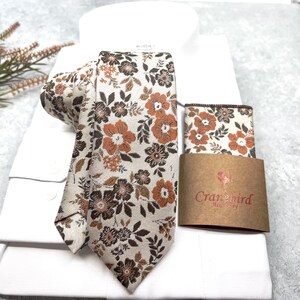 May include: A necktie and matching pocket square set featuring a floral pattern in shades of burnt orange, dark brown, and cream. The set is displayed on a white dress shirt, with a brown paper band labeled Cranebird Accessory.