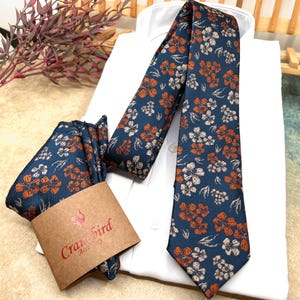 Pode incluir: Uma gravata azul-marinho e um lenço de bolso a condizer com um padrão floral repetitivo em laranja queimado e branco sujo. O lenço de bolso está envolvido numa faixa de papel castanho com o texto vermelho Cranebird Accessory impresso.
