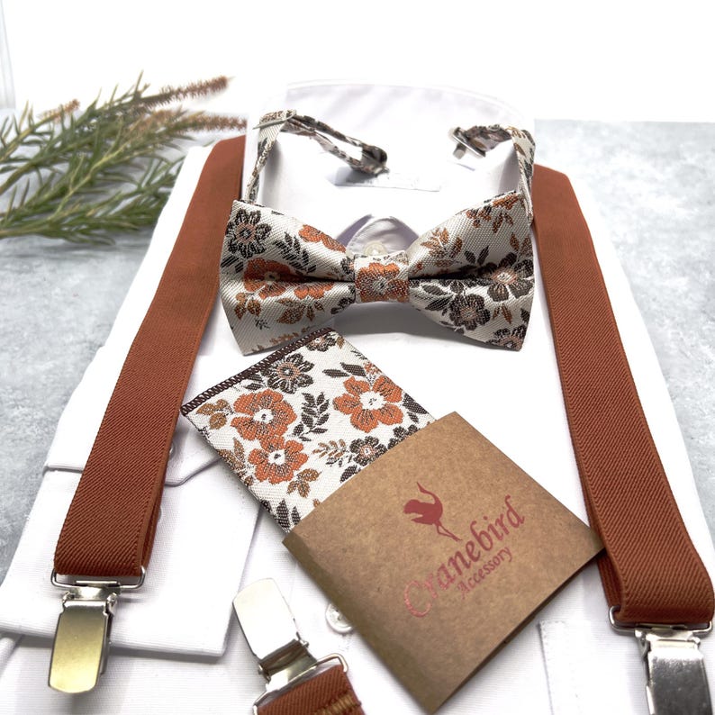 May include: A set featuring a bow tie and pocket square with a white background and a floral pattern in shades of orange and dark brown, paired with solid burnt orange suspenders. A brown paper tag displays the text Cranebird Accessory.