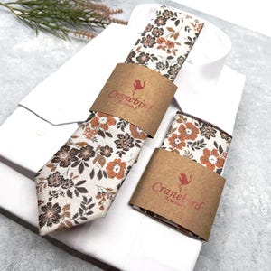 May include: A necktie and a pocket square featuring a floral pattern in shades of brown, orange, and cream. Both items are wrapped in brown paper bands printed with the text Cranebird Accessory, resting on a folded white dress shirt.