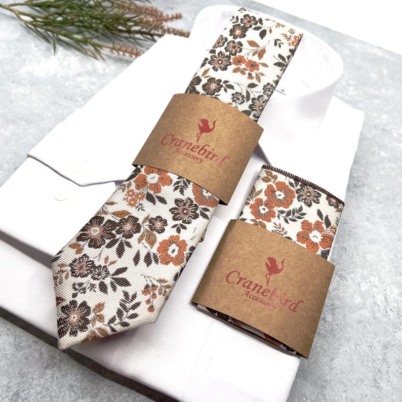 May include: A necktie and a pocket square featuring a floral pattern in shades of brown, orange, and cream. Both items are wrapped in brown paper bands printed with the text Cranebird Accessory, resting on a folded white dress shirt.
