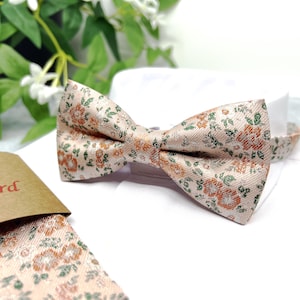 May include: A pre-tied bow tie featuring a light peach background with a repeating pattern of small orange flowers and green leaves. The fabric has a subtle woven texture, and the bow tie is displayed on a white form with a matching fabric piece nearby.