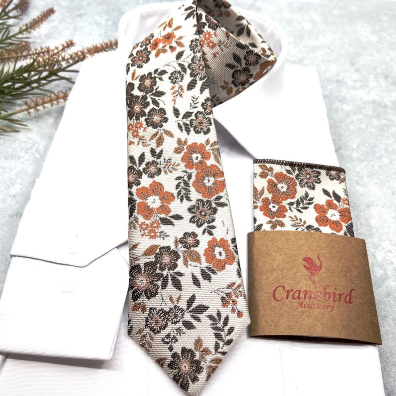 May include: A necktie and matching pocket square featuring a floral pattern in shades of burnt orange, dark brown, and cream. The pocket square is wrapped in a brown paper band with the red text Cranebird Accessory printed on the front.