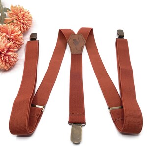 May include: Rust-colored elastic suspenders featuring a central leather patch embossed with an ostrich silhouette. The suspenders include adjustable metal sliders and three antique brass-toned metal clips for attachment to trousers or pants.