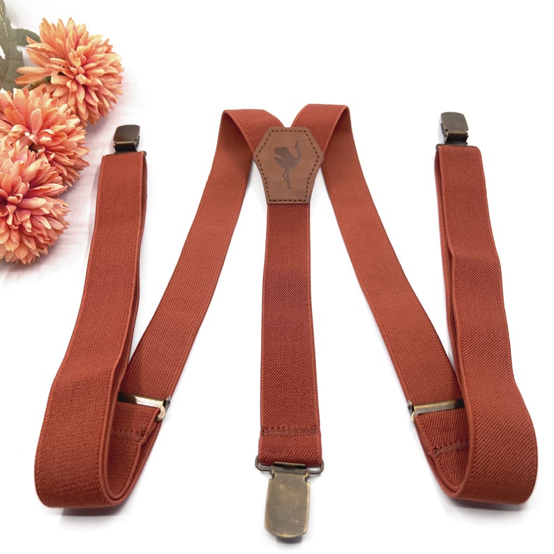May include: Rust-colored elastic suspenders featuring a central leather patch embossed with an ostrich silhouette. The suspenders include adjustable metal sliders and three antique brass-toned metal clips for attachment to trousers or pants.