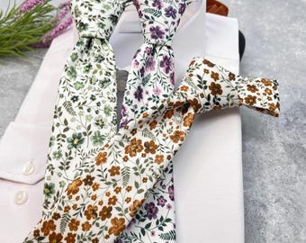 Op de afbeelding: Drie dassen met bloemenpatronen op witte stof. Eén das toont groene bladeren en kleine bloemen, een andere paarse bloesems met groen blad, en de derde bruine bloemen met groene bladeren, allemaal rustend op een wit overhemd.