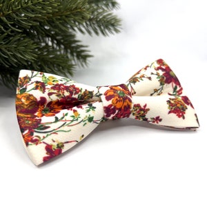 May include: A pre-tied bow tie featuring an off-white fabric base with a vibrant floral pattern in shades of deep red, burnt orange, and dark green. The bow tie has a structured shape with a central knot and a textured, matte finish fabric.
