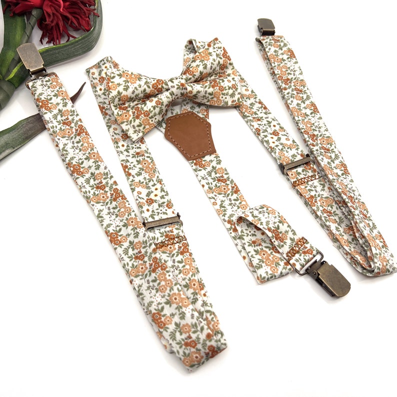May include: A set of matching accessories featuring a floral pattern in white, orange, and green. The set includes a bow tie, suspenders, and a clip-on tie, all with metal hardware and a brown leather accent on the bow tie.