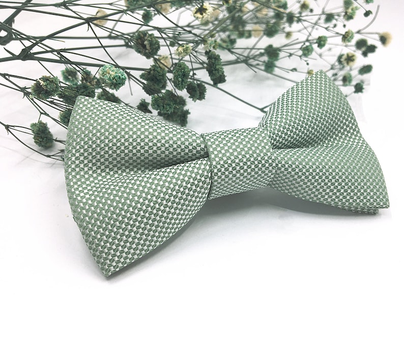 Op de afbeelding: Een saliegroene vlinderdas met een kleinschalig, herhalend geometrisch weefpatroon. De vlinderdas heeft een gestructureerde, symmetrische vorm met een centrale knoop en twee puntige vleugels, met een getextureerde stoffen afwerking in groen.