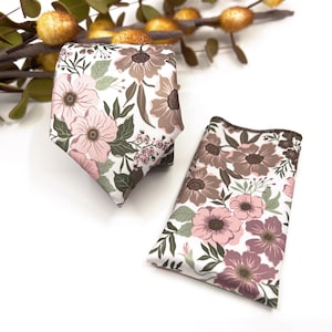 Peut inclure: Un ensemble de cravate et de pochette de poche à motif floral dans des tons de rose, marron et blanc. La cravate a une extrémité pointue et la pochette de poche est carrée.