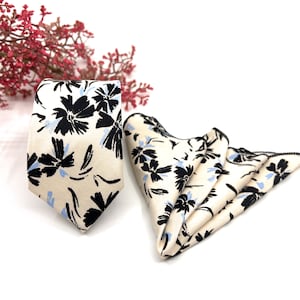 May include: A necktie and a folded pocket square featuring a cream-coloured base with a printed pattern of black and light blue floral motifs. The fabric has a smooth, satin-like finish, and the items are displayed together on a plain surface.