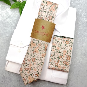 May include: A necktie and a matching pocket square featuring a floral pattern in shades of peach, brown, and green on a light background. A brown paper band with the text Cranebird is wrapped around the necktie, which rests on a white shirt.