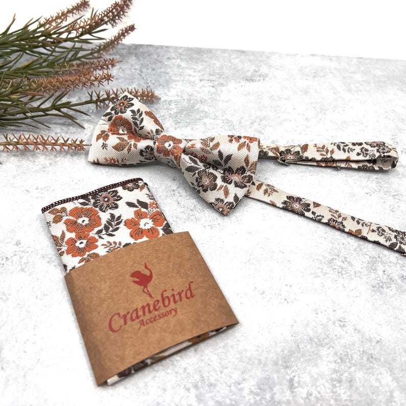 May include: A bow tie and a pocket square featuring a floral pattern in shades of orange, brown, and cream. The pocket square is partially wrapped in a brown paper band with the text Cranebird Accessory and a small bird logo printed in red.