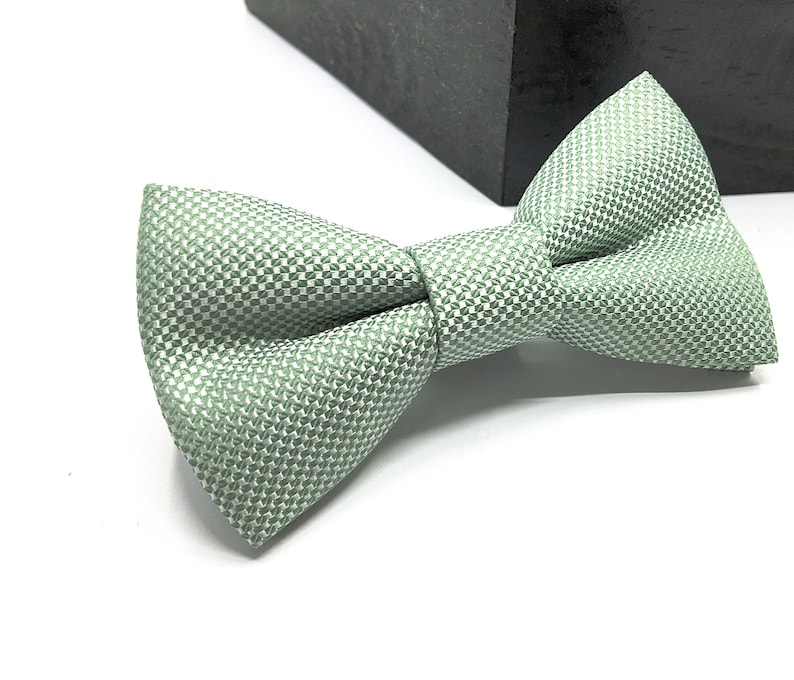 Op de afbeelding: Een saliegroene vlinderdas met een fijn, herhalend geometrisch weefpatroon. Het accessoire heeft een gestructureerde, voorgeknoopte vorm met een centraal knoopdetail en een textieloppervlak dat een subtiel lichtreflecterend effect geeft.