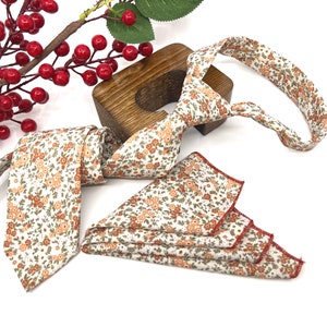 May include: A necktie and a matching pocket square featuring a white fabric base with a repeating floral pattern in shades of orange, brown, and green. The pocket square has a finished edge with red stitching, and the necktie has a pointed end.