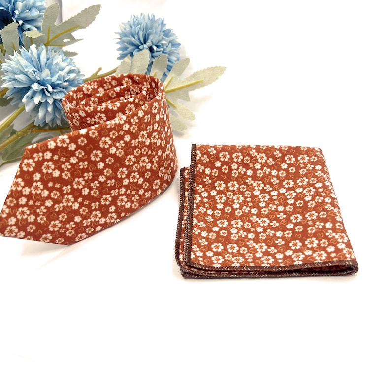 May include: A necktie and a matching square pocket handkerchief, both featuring a rust-colored fabric with a repeating pattern of small white flowers. The items are displayed alongside light blue artificial flowers with green stems.