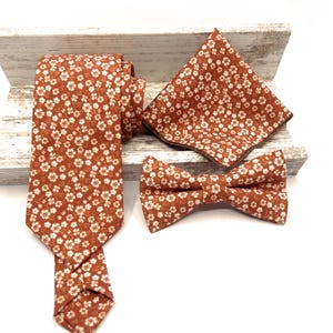 May include: A set consisting of a necktie, a bow tie, and a pocket square, all featuring a matching burnt orange fabric with a repeating pattern of small white flowers. The items are arranged together to display the consistent floral print.