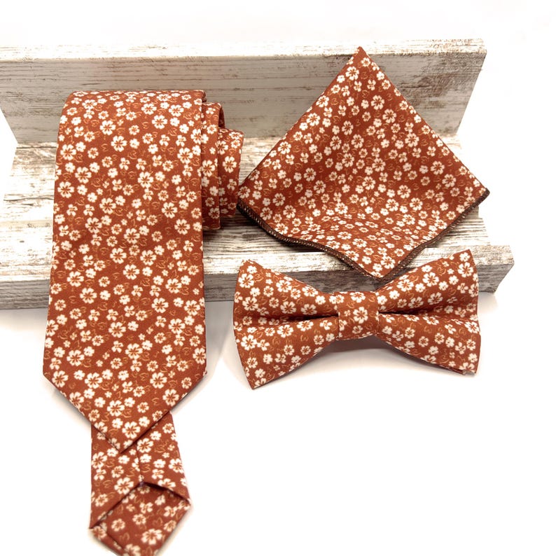 May include: A set consisting of a necktie, a bow tie, and a pocket square, all featuring a matching burnt orange fabric with a repeating pattern of small white flowers. The items are arranged together to display the consistent floral print.