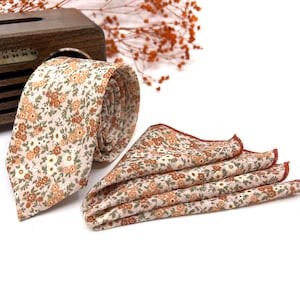 Op de afbeelding: Een stropdas en een gevouwen pochet met een herhalend bloemenpatroon van kleine oranje, bruine en witte bloemen op een lichtbeige achtergrond. Het pochet heeft een afgewerkte roestkleurige gestikte rand langs de driehoekige vouwen.