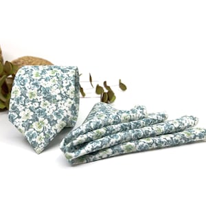 Puede incluir: Una corbata y un pañuelo de bolsillo doblado con un estampado floral en tonos verde salvia, verde azulado y blanco. La tela muestra una disposición densa de pequeñas flores superpuestas en toda la superficie de ambos artículos.