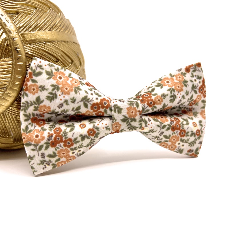 May include: A floral patterned bow tie with a white background and small orange and green flowers. The bow tie is made of fabric and is tied in a classic bow shape. The bow tie is a fashion accessory.