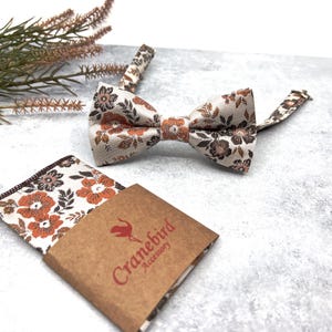 May include: A bow tie and a matching pocket square featuring a floral pattern in shades of orange, brown, and cream. The pocket square is partially wrapped in a brown paper band with the red text Cranebird Accessory printed on the front.