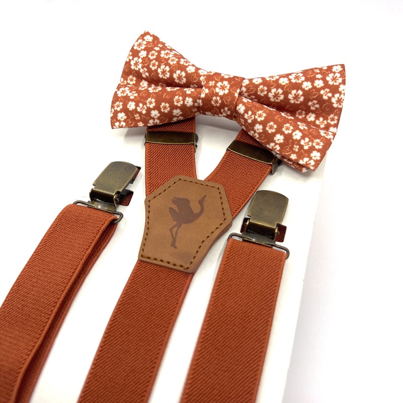May include: A set consisting of a bow tie and suspenders in a burnt orange color. The bow tie features a small white floral pattern. The suspenders have solid orange elastic straps, bronze-toned metal clips, and a central leather-like patch.