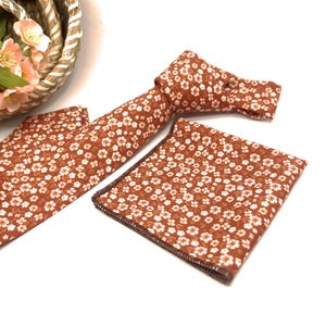 May include: A rust-colored necktie and a matching square pocket handkerchief, both featuring a repeating pattern of small white five-petaled flowers. The necktie is tied in a knot, and the pocket square has a dark brown stitched edge.