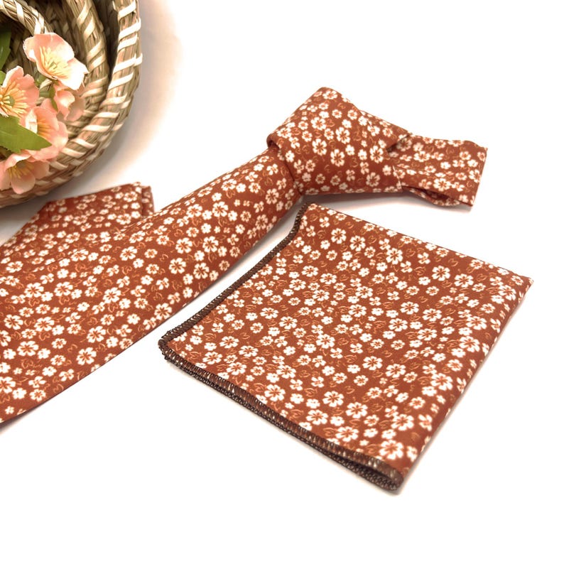 May include: A rust-colored necktie and a matching square pocket handkerchief, both featuring a repeating pattern of small white five-petaled flowers. The necktie is tied in a knot, and the pocket square has a dark brown stitched edge.